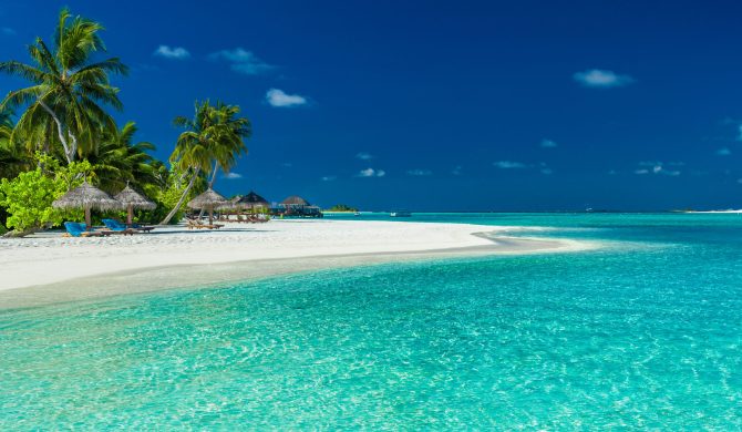 Palm trees and beach umbrelllas over lagoon and white sandy beach, Maldives island