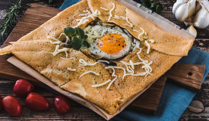 French pancake crepe with fried egg, herbes, tomatoes and cheese on a slat on blue linen on a wooden table. Closeup.