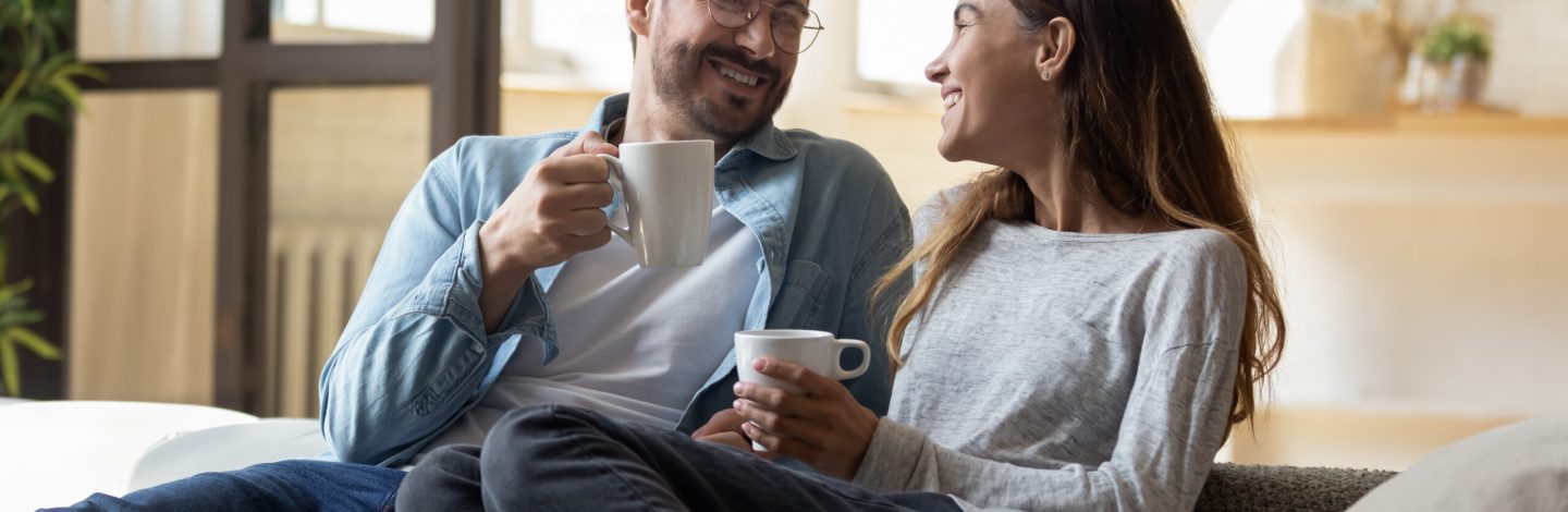 Calm relaxed couple hold cups rest on comfy sofa enjoy carefree talk while sit together on couch at modern living room. Homeowners family spend weekend at home, morning beverage, communication concept