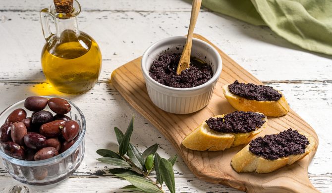 Bread or crostini with olive pate on wooden cutting board. Olives, oil, green napkin and olive branch aside on white wooden background
