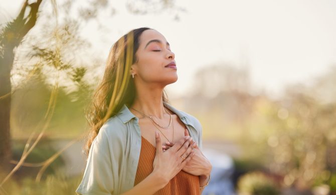 Young latin woman with hand on chest breathing in fresh air in a beautiful garden during sunset. Healthy mexican girl enjoying nature while meditating during morning exercise routine with closed eyes. Mindfulness woman enjoying morning ritual and relaxing technique.